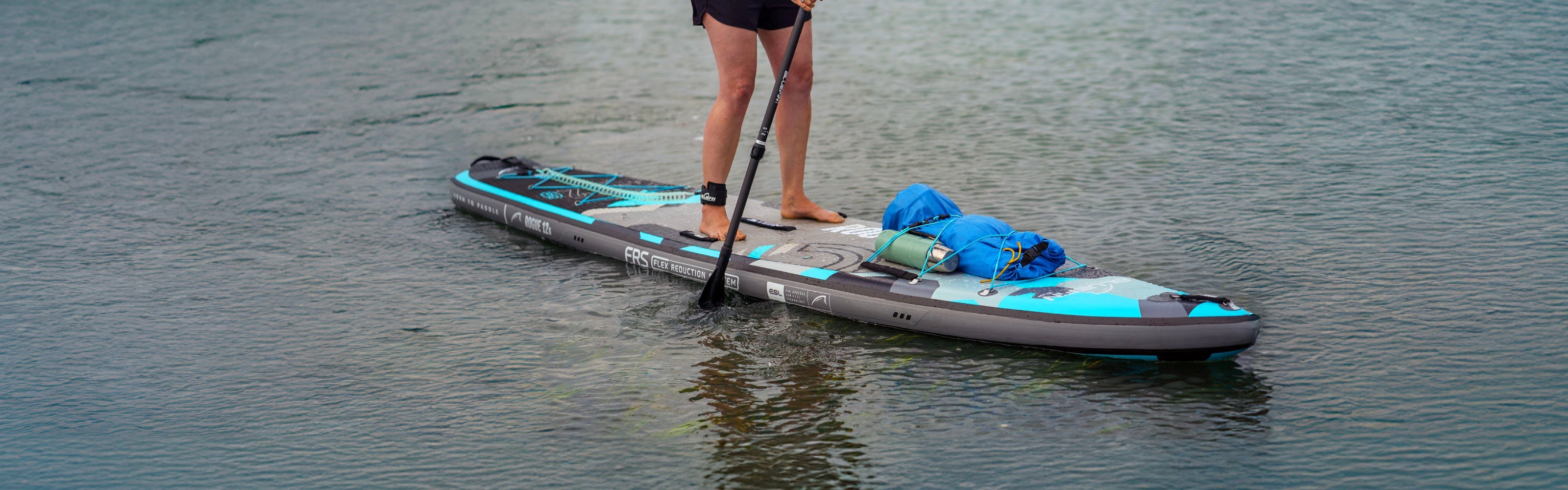 Person standing on a paddleboard with a blue bag, paddling on calm water