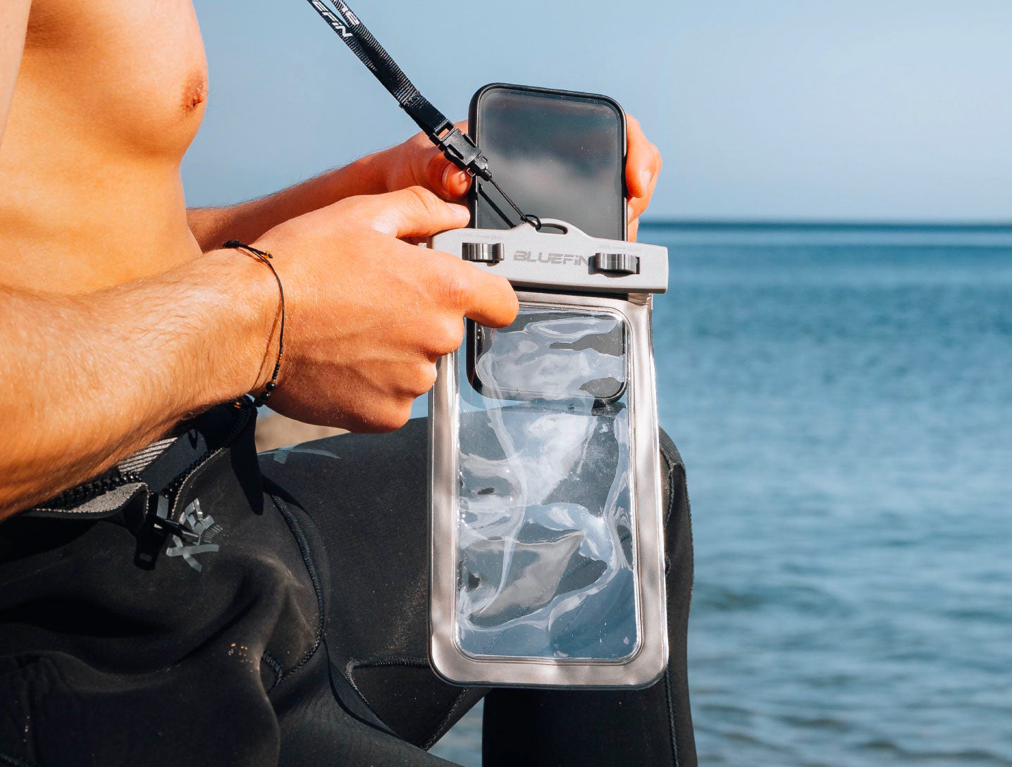 Person holding a waterproof phone case by Bluefin near the water, wearing a wetsuit.