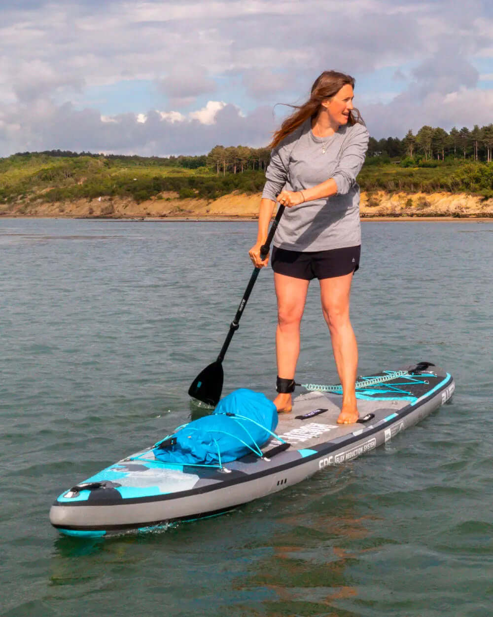 Woman paddling on an inflatable paddleboard with a blue bag, wearing a gray long-sleeve shirt and shorts.