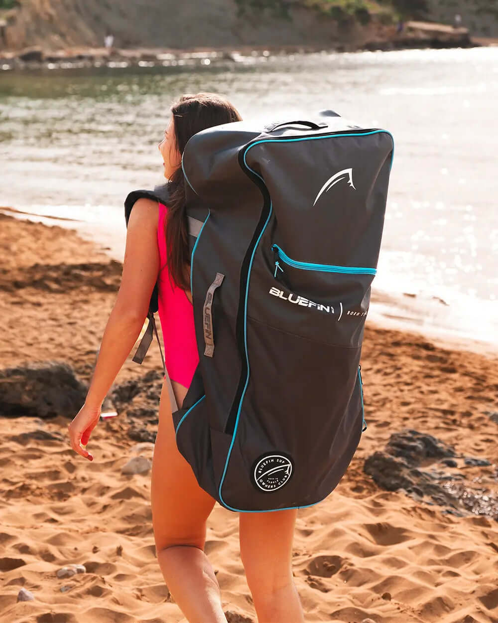 Woman in a pink swimsuit carrying a large black Bluefin SUP backpack on a sandy beach.