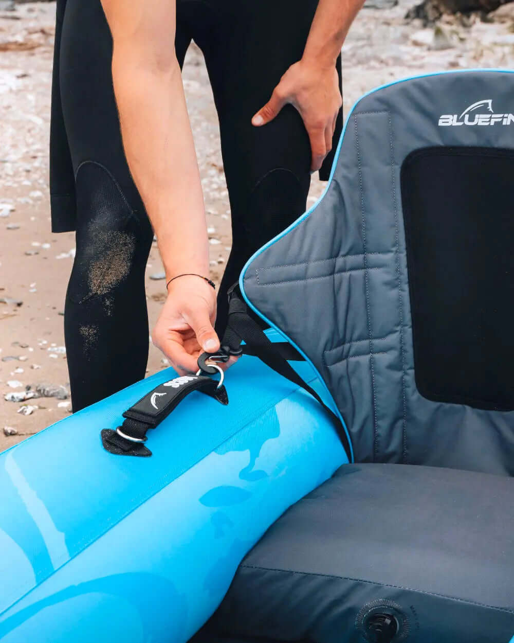 Person in wetsuit adjusting strap on blue inflatable paddleboard on sandy beach