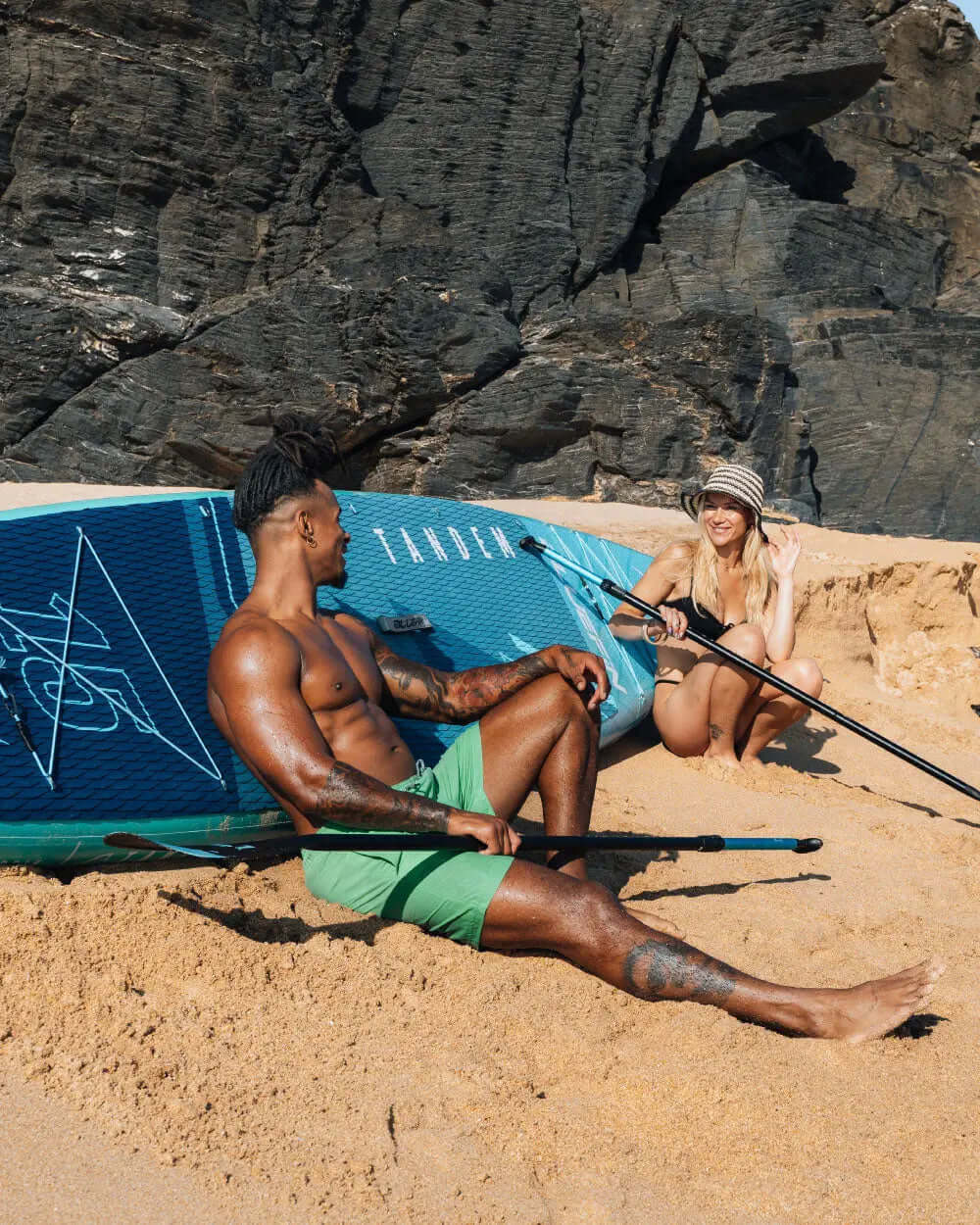 Two people sitting on a sandy beach with a paddleboard, one wearing green shorts and the other in a swimsuit.