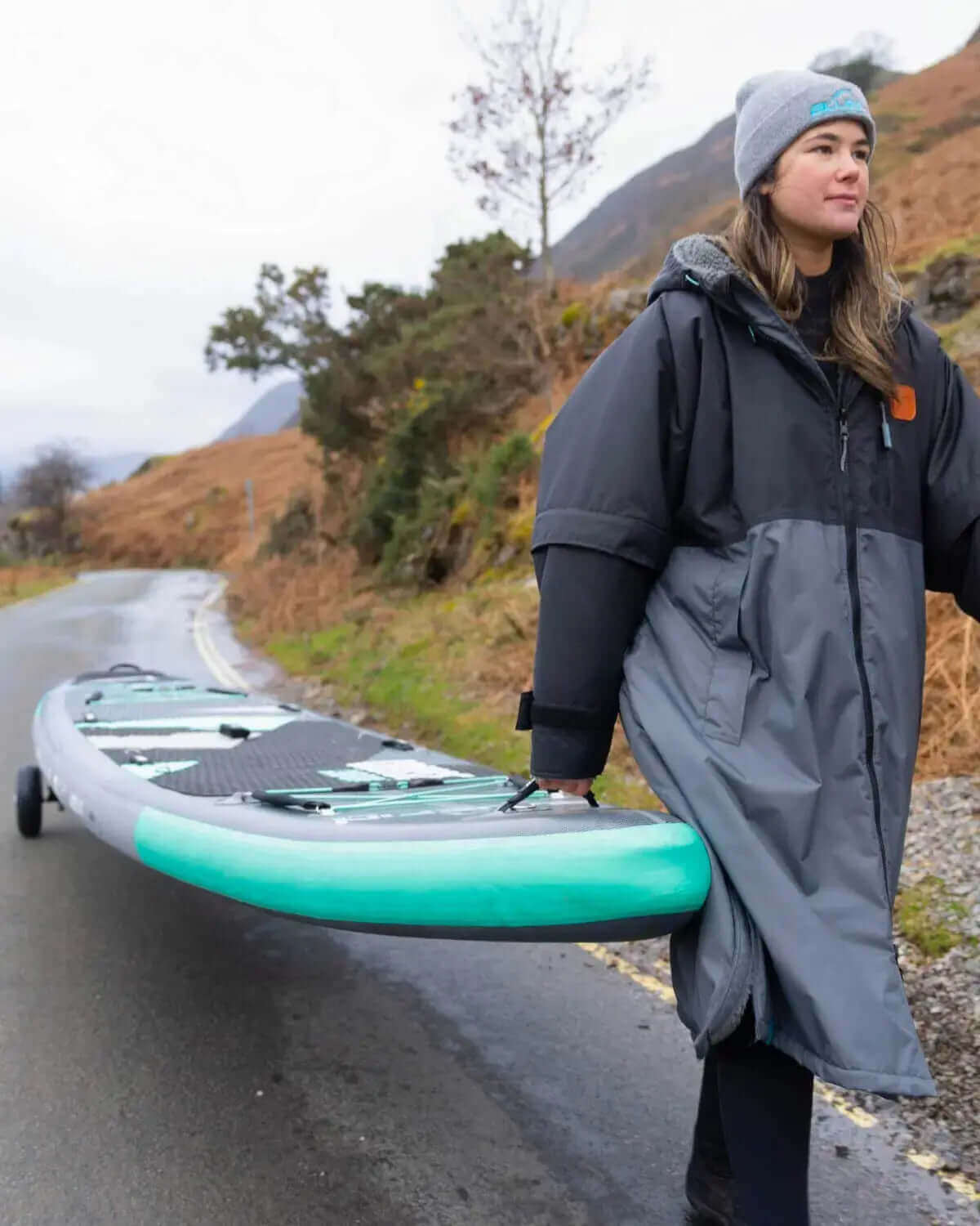 Pers in een grijze jas en beanie die een teal opblaasbare paddleboard langs een weg draagt.