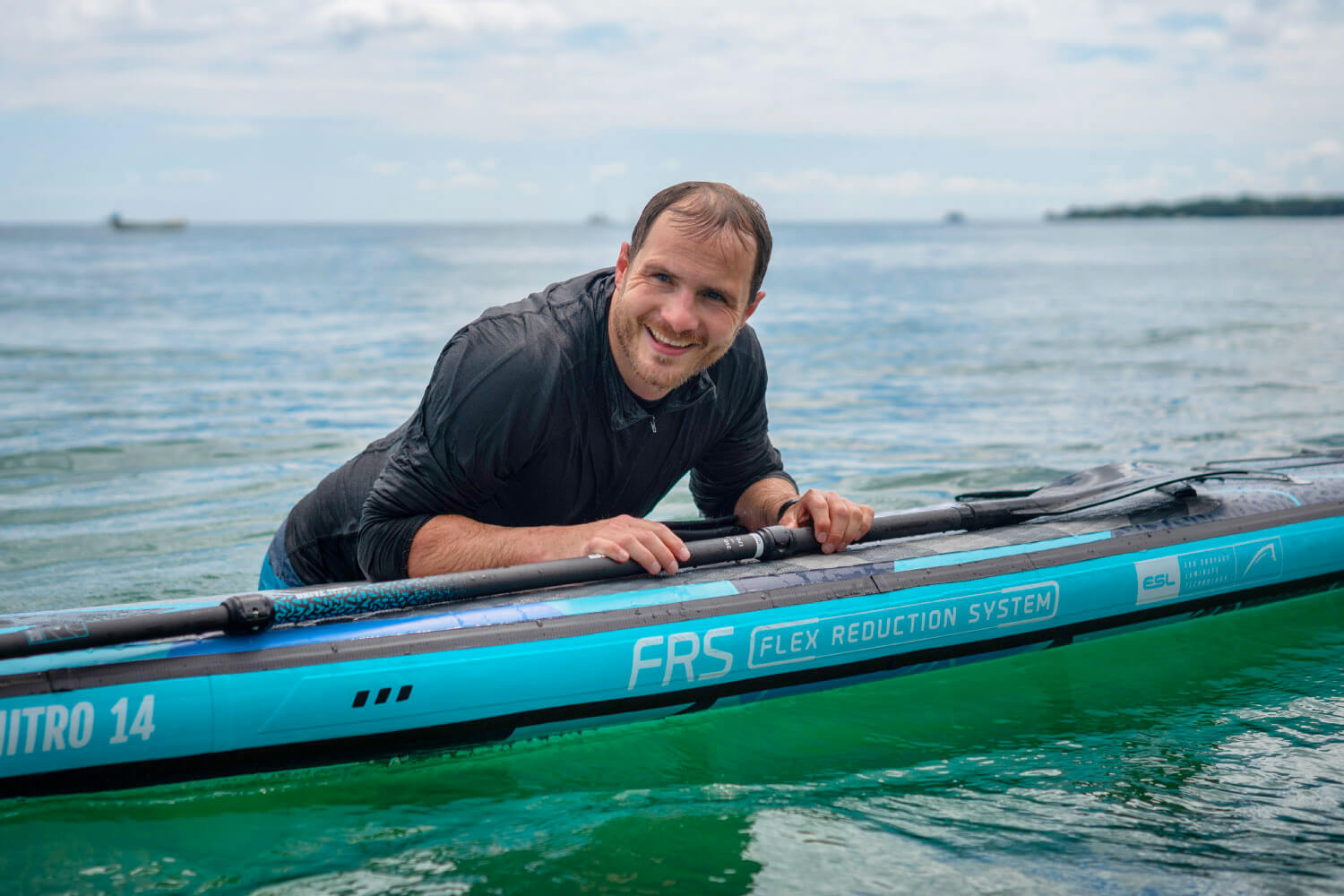Man smiling while leaning on an inflatable paddleboard in calm water, with FRS branding visible