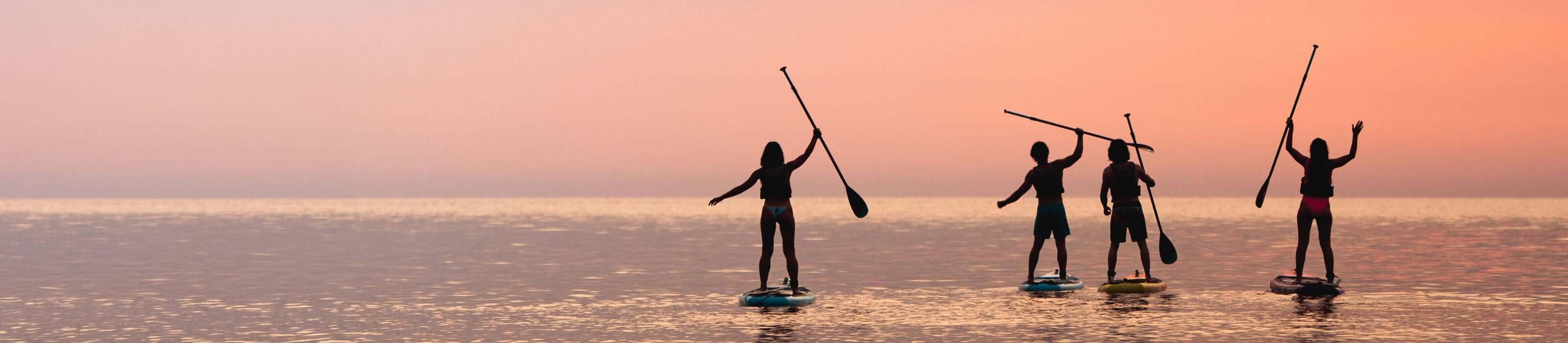Four people paddleboarding at sunset, silhouetted against a pink sky on calm water.