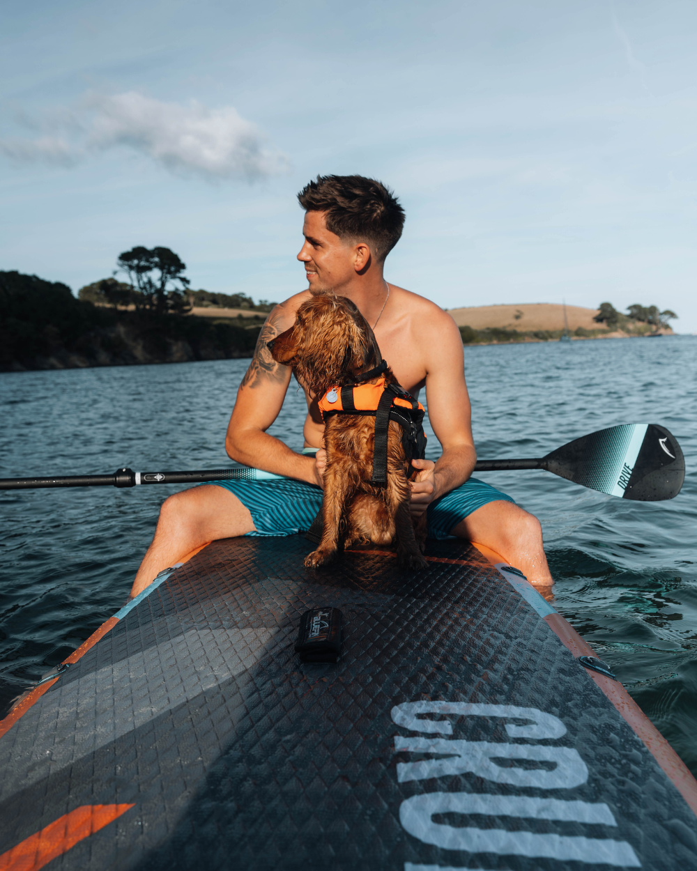Man on a paddleboard with a dog, both enjoying a calm water setting, wearing life jackets.