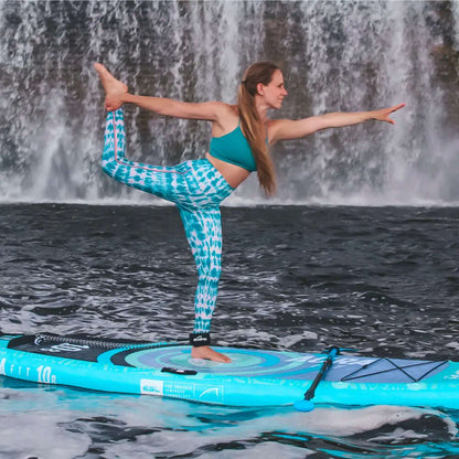 Frau in tealfarbener Sportbekleidung, die Yoga auf einem Paddleboard vor einem Wasserfall macht