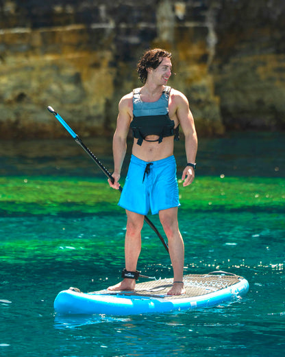 Man on a blue inflatable paddleboard, wearing a life vest and blue shorts, paddling in clear water.