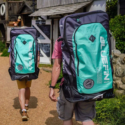 Deux personnes marchant avec de grands sacs à dos Bluefin Carbon SUP sur un chemin de gravier près de bâtiments.