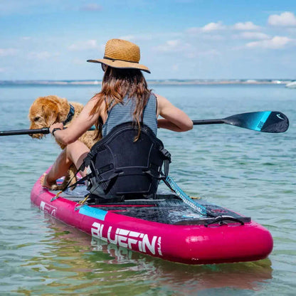 Persona remando en una tabla de paddle inflable rosa con un perro, usando un sombrero de paja, en agua clara.