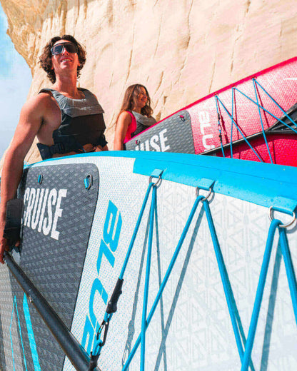 Two people carrying inflatable paddleboards with blue and red designs near a rocky cliff.