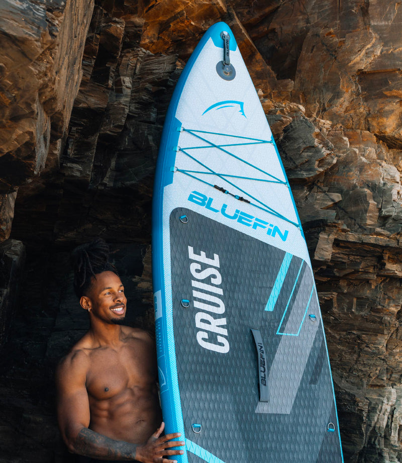 Inflatable Bluefin paddleboard with textured deck and center fin, man smiling beside rocky background