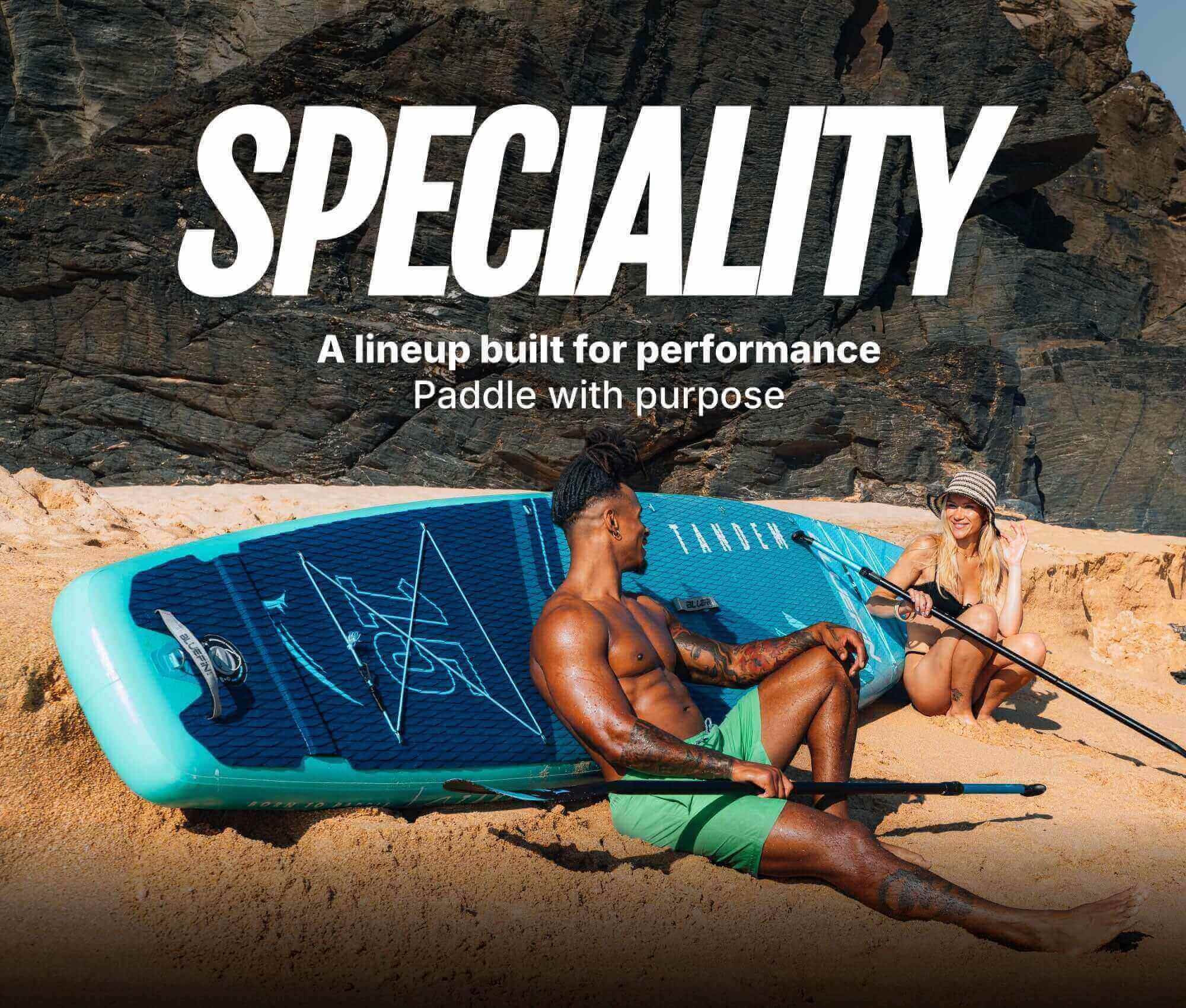 Two people on a sandy beach with an inflatable paddleboard, one sitting and one waving, both with paddles.