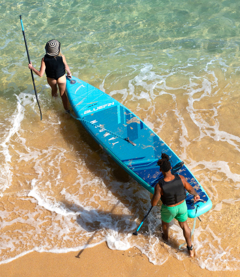 Two people walking into the water with a blue inflatable paddleboard and paddles, sandy beach in background