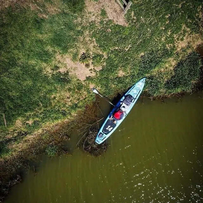 Vista aerea di un kayak blu su una riva verde con una persona che pagaia e erba che circonda l'acqua.