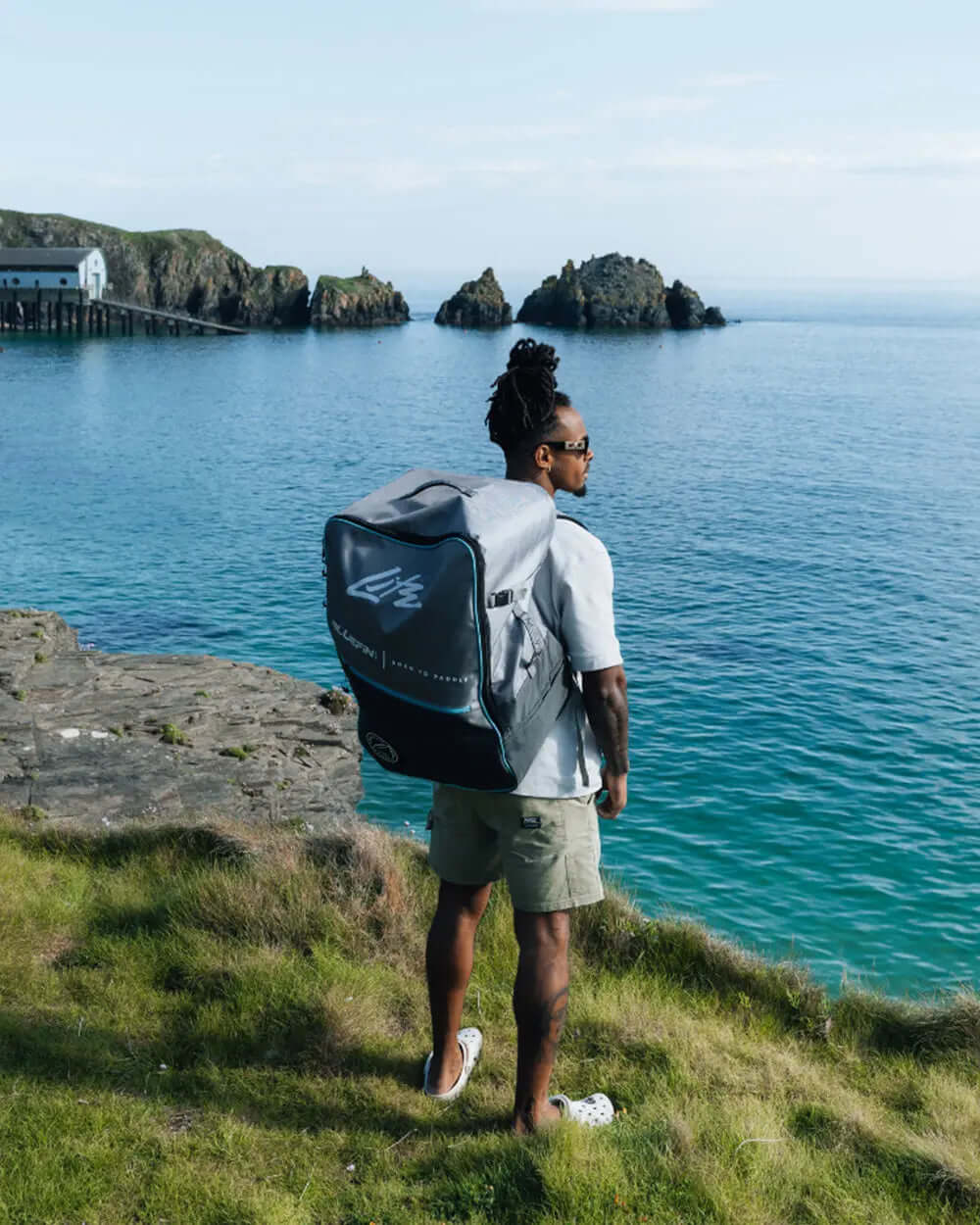 Person wearing sunglasses and a backpack stands on a grassy cliff overlooking the ocean and rocky islands.
