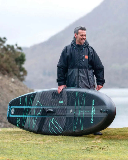 Man in black jacket standing on grass with a Bluefin inflatable paddleboard, mountains in background