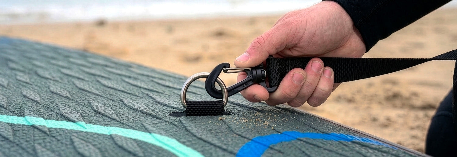 Hand attaching a strap to a D-ring on a textured paddleboard at the beach
