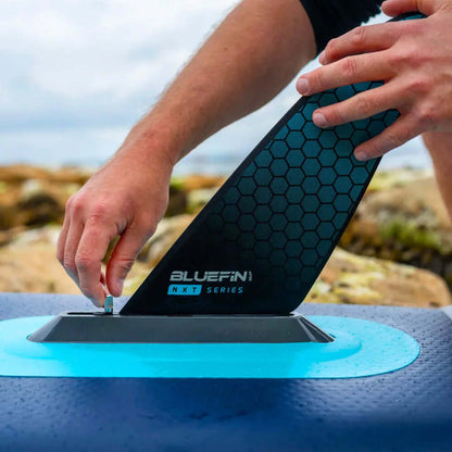 Personne fixant un aileron hexagonal noir à une planche de paddle Bluefin sur une côte rocheuse.