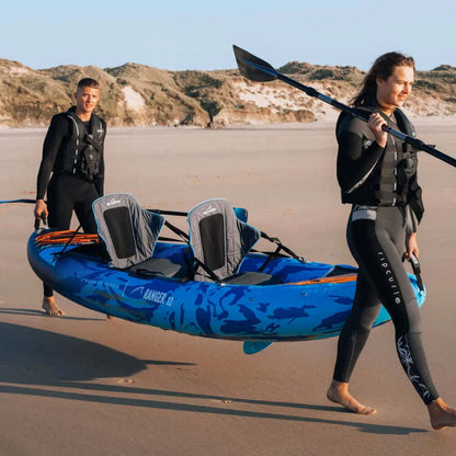 Dos personas caminando por la playa con un kayak inflable azul, una llevando un remo.