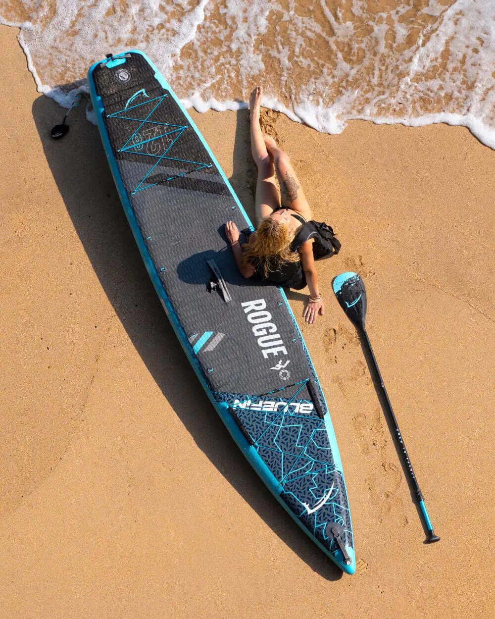 A woman lounging on a blue and black inflatable paddleboard on sandy beach, paddle beside her.