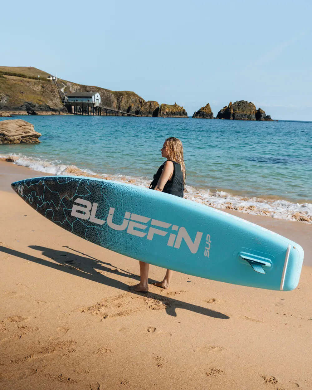 Person carrying a Bluefin SUP paddleboard on a sandy beach near the water