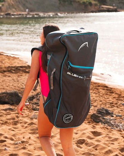 Woman in a pink swimsuit carrying a large black Bluefin SUP backpack on a sandy beach.