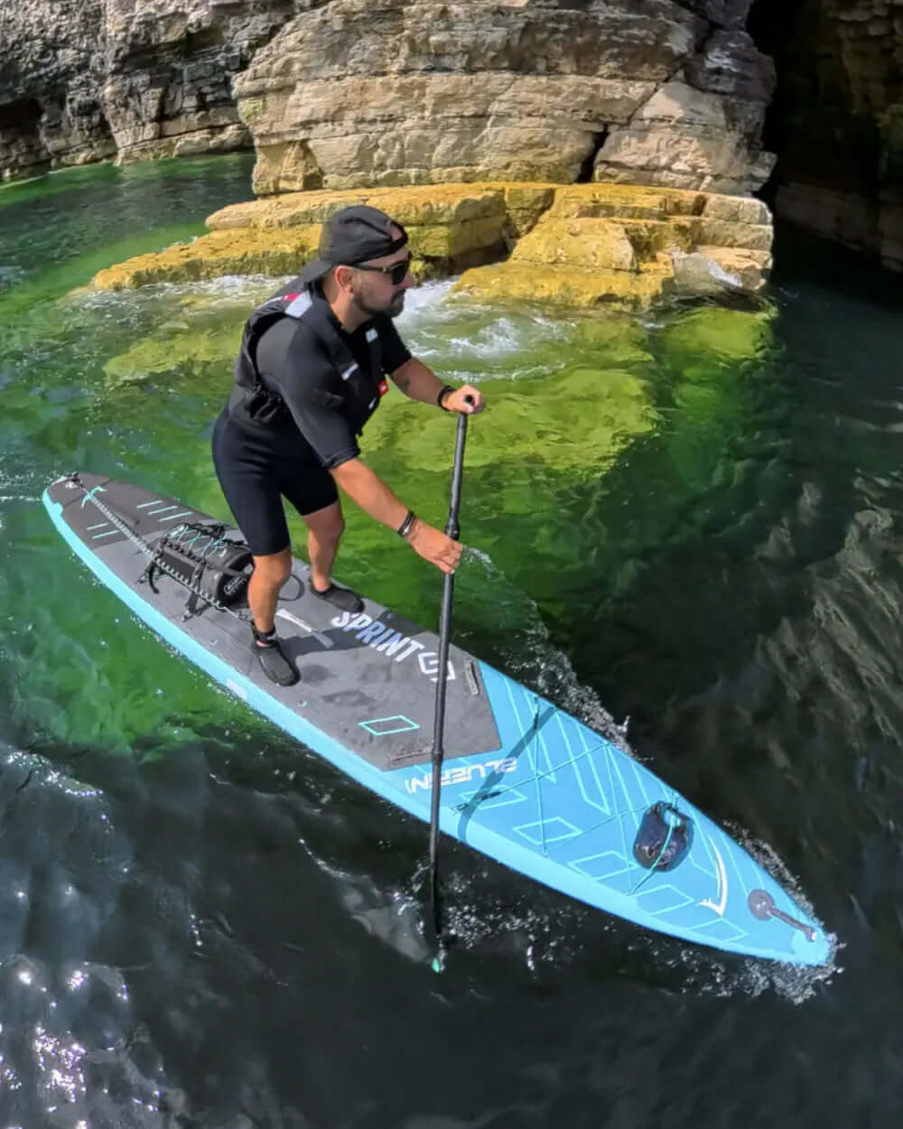 Man paddling on a blue inflatable paddleboard in clear green water near rocky cliffs