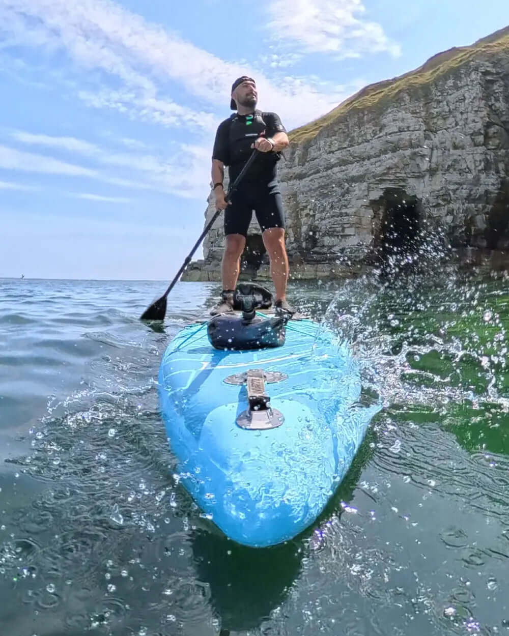 Paddleboarder on a blue inflatable paddleboard, paddling in calm water near rocky cliffs.