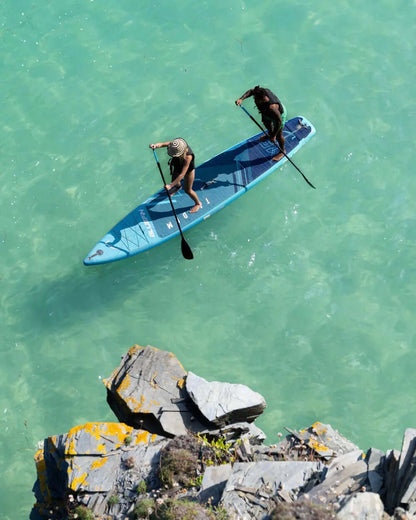 Two people paddling on a blue stand-up paddleboard in clear water near rocky shore
