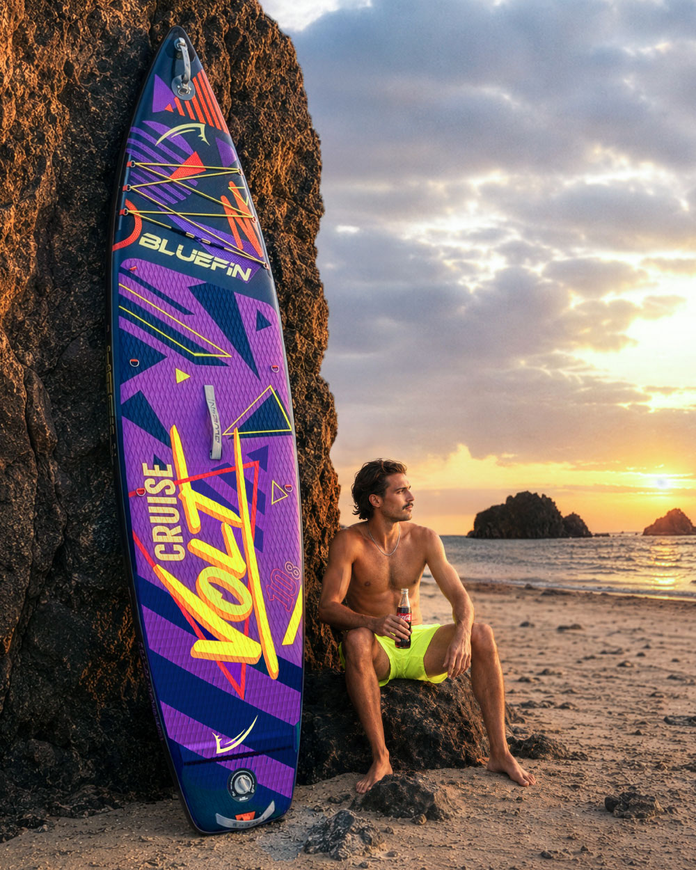 Inflatable paddleboard with colorful design, man in yellow shorts sitting on beach at sunset