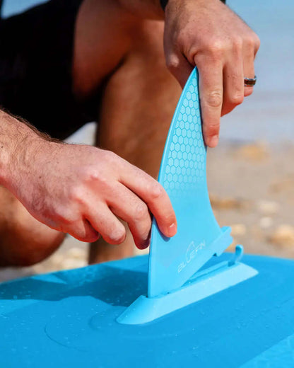 Person attaching a blue fin to an inflatable paddleboard on a sandy beach